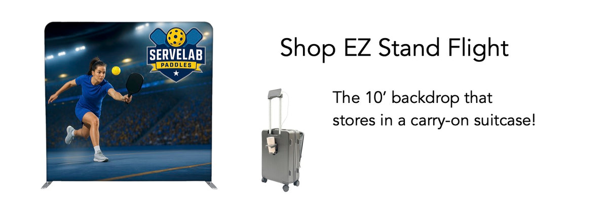 A promotional banner for an EZ Stand flight-sized backdrop, showing a printed tennis-themed backdrop stand on the left and a gray carry-on suitcase on the right with text reading 'Shop EZ Stand Flight' and 'The 10' backdrop that stores in a carry-on suitcase!'.
