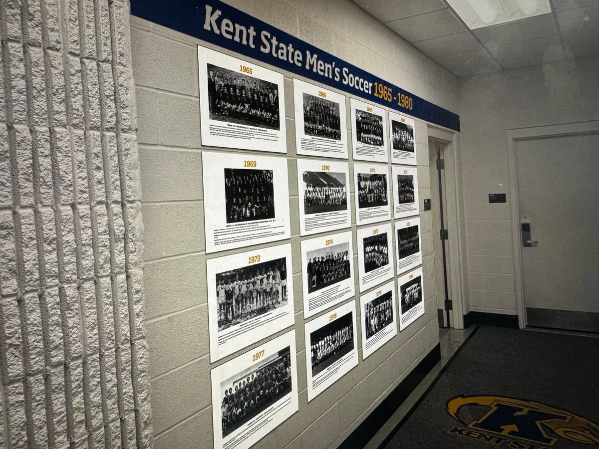 A hallway wall display featuring multiple black-and-white historical team photos printed 3mm PVC Sintra under a blue BrickWrap sticker that reads 'Kent State Men's Soccer 1965-1990.'. In Kent, OH